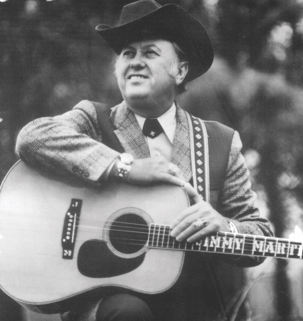Portrait of Jimmy Martin smiling with his guitar resting on his knee.