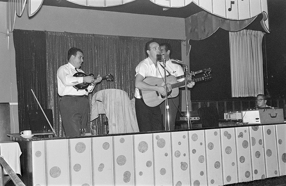 The Osborne Brothers with Benny Birchfield at Ruby’s White Sands outside Dayton, Ohio on May 28, 1964.  Sandy Rothman, seated, is visible taping the show to the right. Jerry was there but is not in the frame. L-R:  Bobby Osborne, Benny Birchfield, and Sonny Osborne. 
Photo by Neil Rosenberg, courtesy of Sandy Rothman.